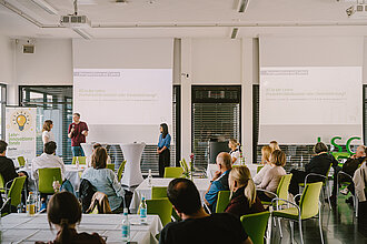 Drei Personen stehen beim Tag der Lehre 2026 vor Publikum in einem hellen Seminarraum mit grünen Stühlen.