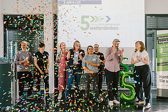 Gruppe von acht Personen steht vor einer Wand mit Schriftzug '5 Jahre Lehre gemeinsam weiterdenken', bunte Konfetti fällt, große grüne Ballonzahl '5' steht rechts