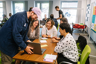 Einige Menschen sitzen im Rahmen eines Workshops an einem Tisch und schauen gemeinsam auf einen Laptop