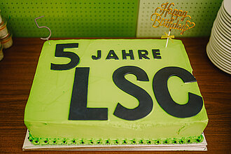 Rechteckige grüne Geburtstagstorte mit schwarzer Aufschrift '5 JAHRE LSC' und einem goldfarbenen 'Happy Birthday'-Stecker auf einem Holztisch neben einem Stapel weißer Teller.
